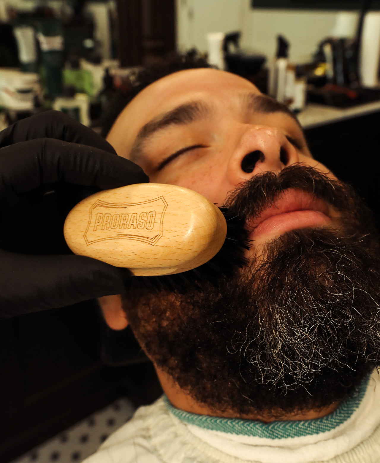 Man with a thick beard having it brushed by a barber with a barbershop in the background