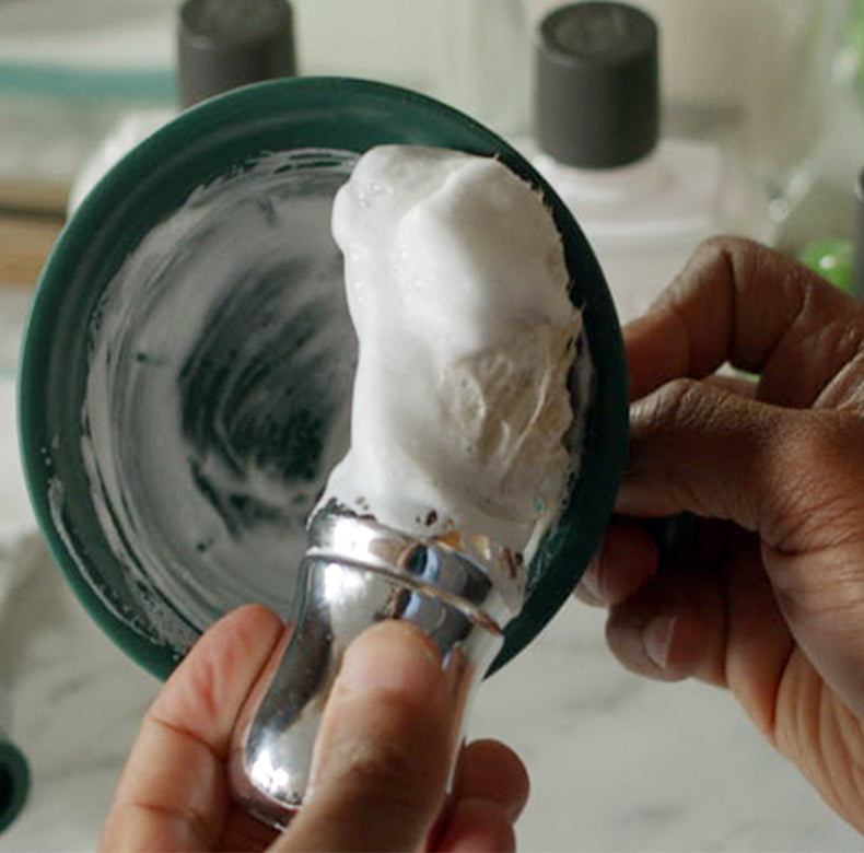 Barber making a thick lather in a green shave mug with After Shave Lotion bottles in the background