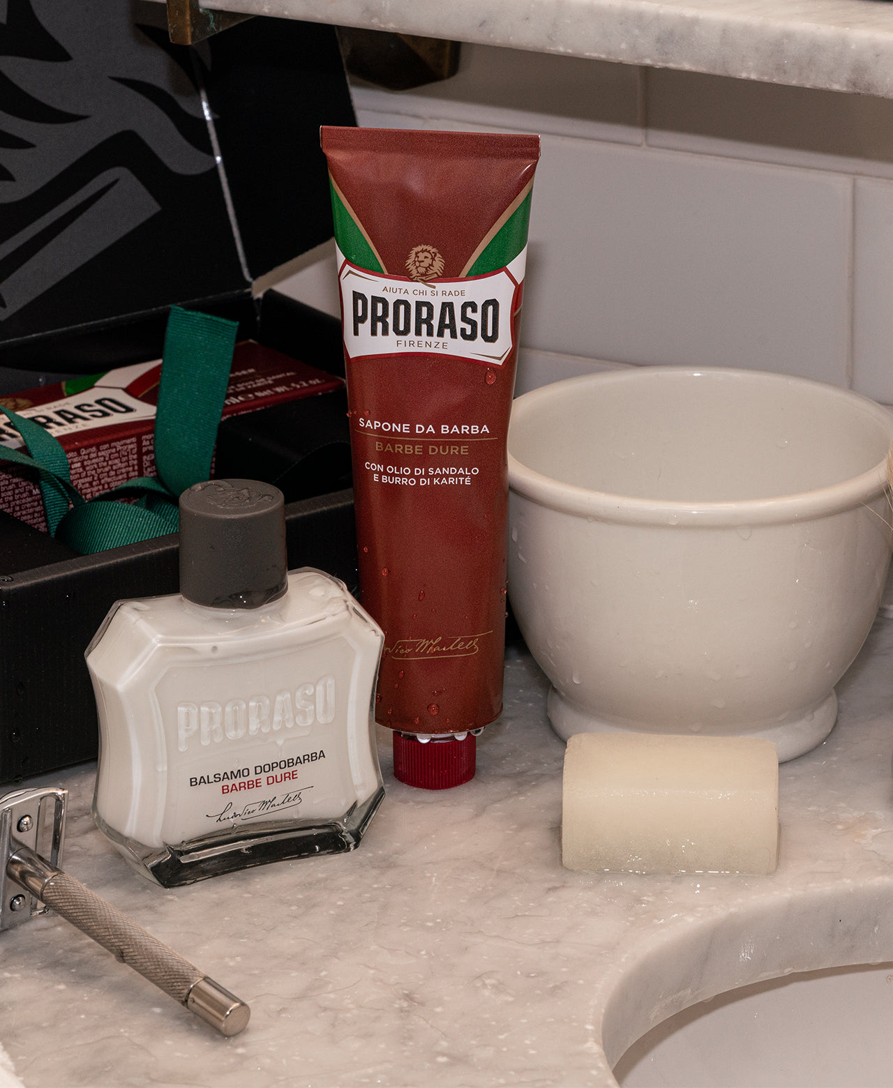 Components of Nourishing Classic Shaving Duo Box on a white marble counter After Shave Balm and Shave Cream Tube with a silver double edge safety razor on the left, Post-Shave Stone and shave mug on the right with Duo Box in the background