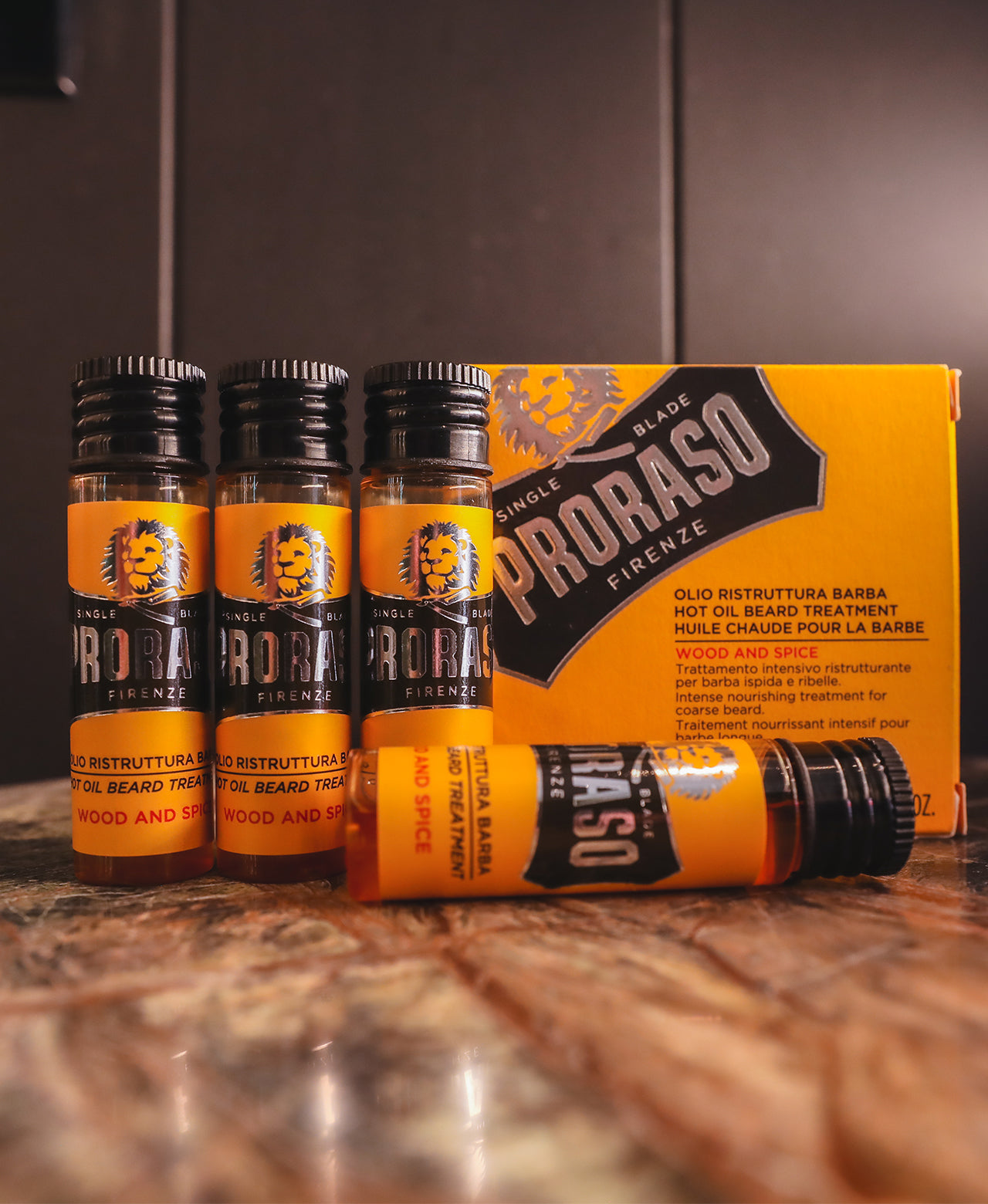 Three Hot Oil Beard Treatment vials standing up next to product box with one vial laying on it's side in front, on a brown marble table with a black wooden background