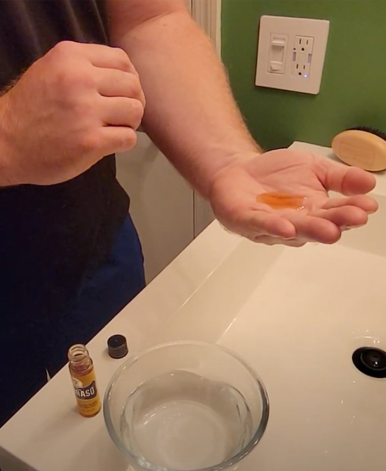 Vial of Hot Oil Beard Treatment poured into a man's palm standing in front of bathroom sink ready to apply to beard