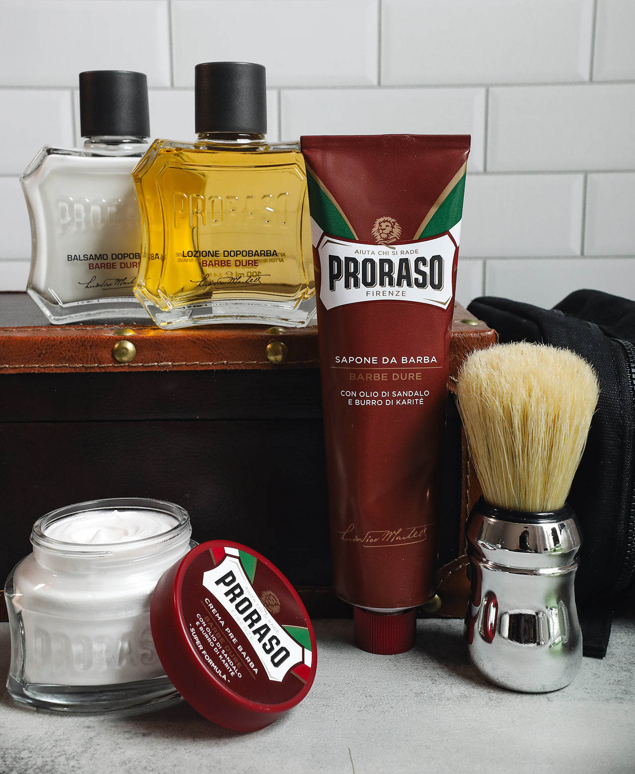 Coarse Beard Shave formula lineup: Pre-Shave Cream with open lid, Shave Cream Tube and Professional Shaving Brush in front. After Shave Balm and After Shave Lotion sit in the background on top of a vintage suitcase with a black dopp kit all in front of a white subway tile background 