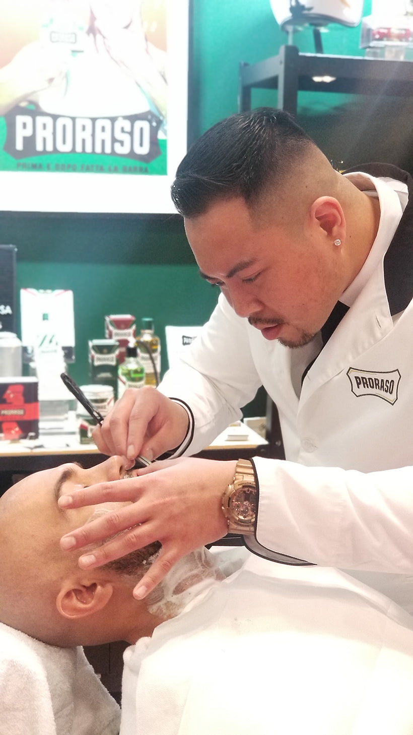 Barber shaving the edges of a mans beard who has shaving cream on his face reclined in a barber chair
