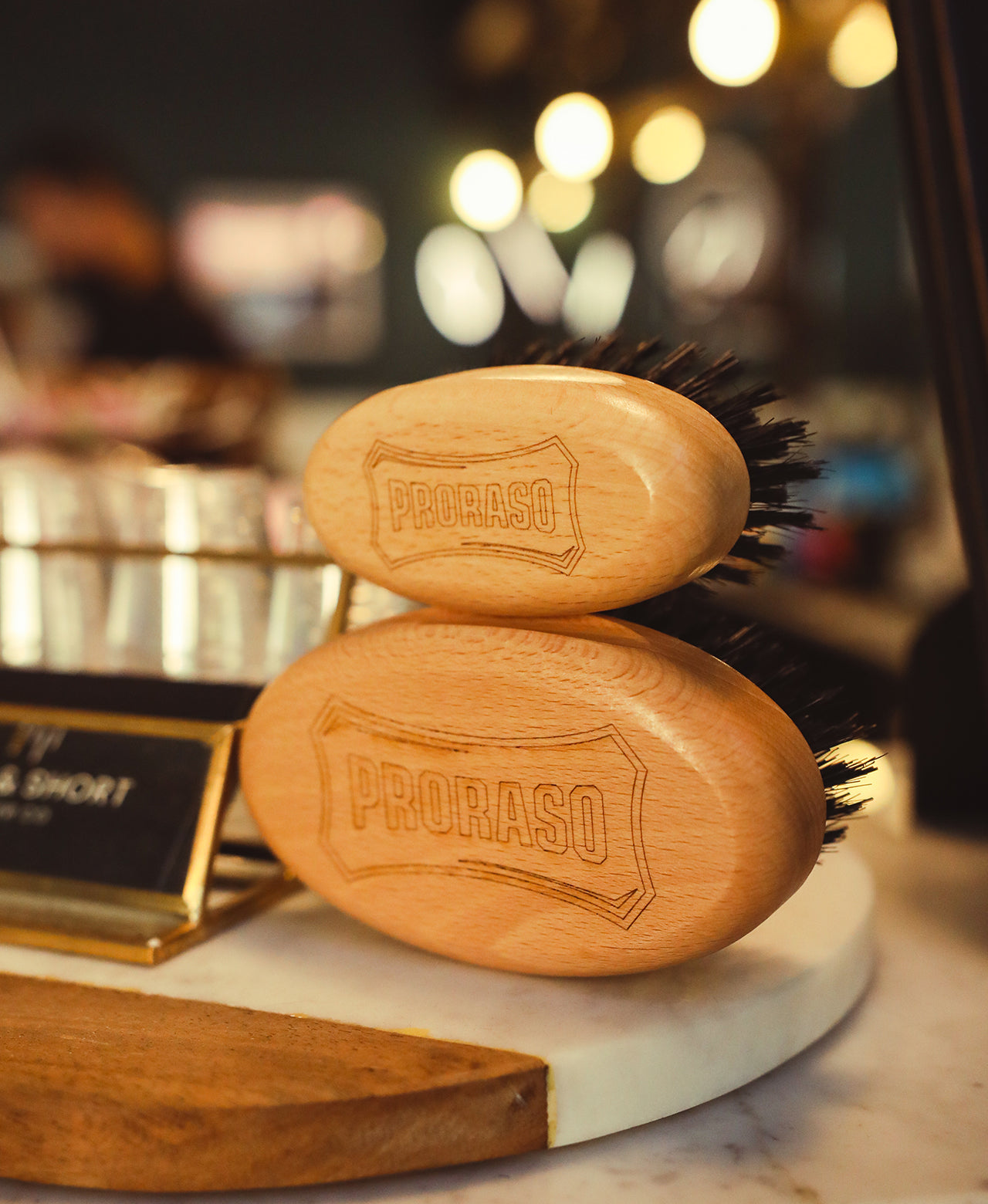 An image to show scale, the smaller Moustache and Beard Brush sitting on top of the Old Style Military Brush sitting on the marble counter at a barbershop with the background obscured 
