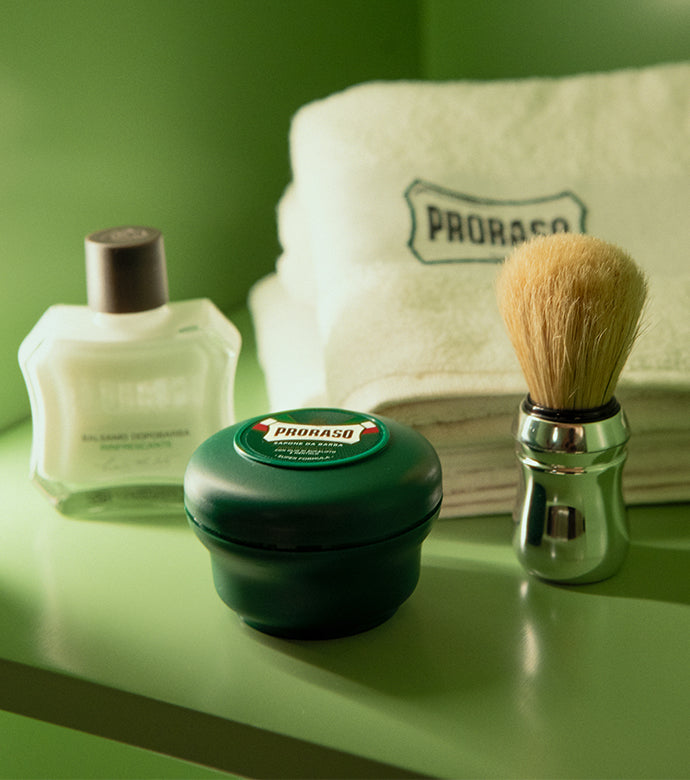 Refreshing products from left to right After Shave Balm, Shaving Soap in a Bowl and Professional Shave Brush with white Proraso barber towels in the background sitting in green shelves