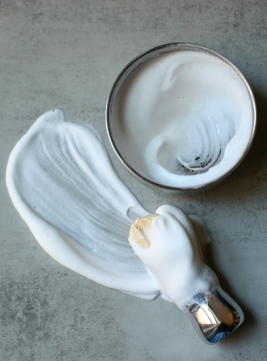 Our Professional Shave Brush with a thick lather on it laying on a grey table with a silver bowl full of lather next to it