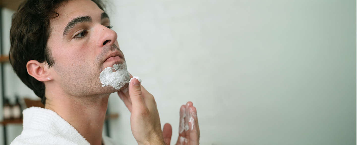Young man applying Pre-Shave Cream to his chin on bathroom wearing white robe