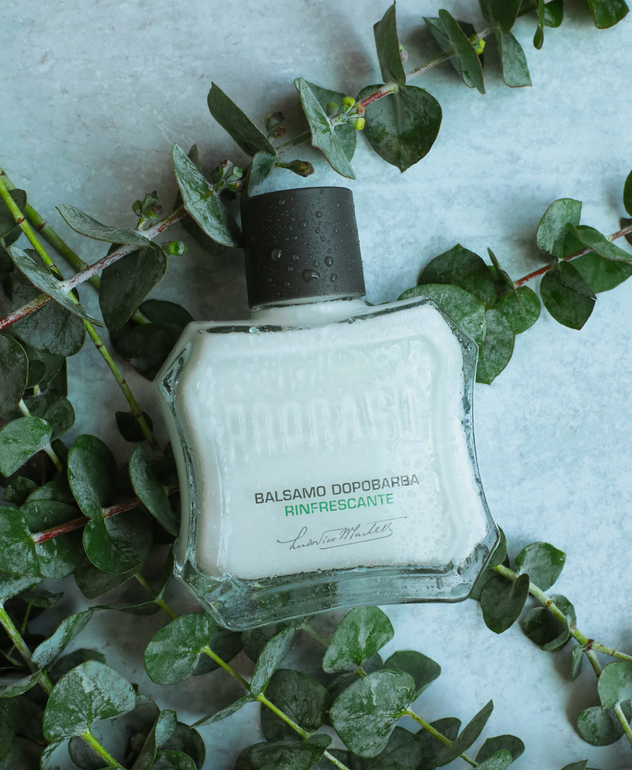 Bottle of Refreshing After Shave Balm on eucalyptus leaves on a grey table 