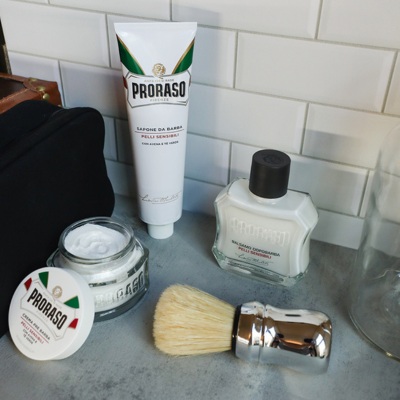Sensitive formula Pre-Shave Cream, Shave Cream Tube and After Shave Balm with a Professional Shave Brush sitting in front of a suitcase and white tile background